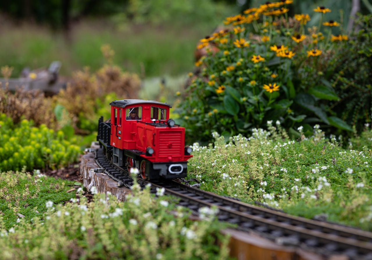A vibrant red toy train traverses a green garden track surrounded by flowers.
